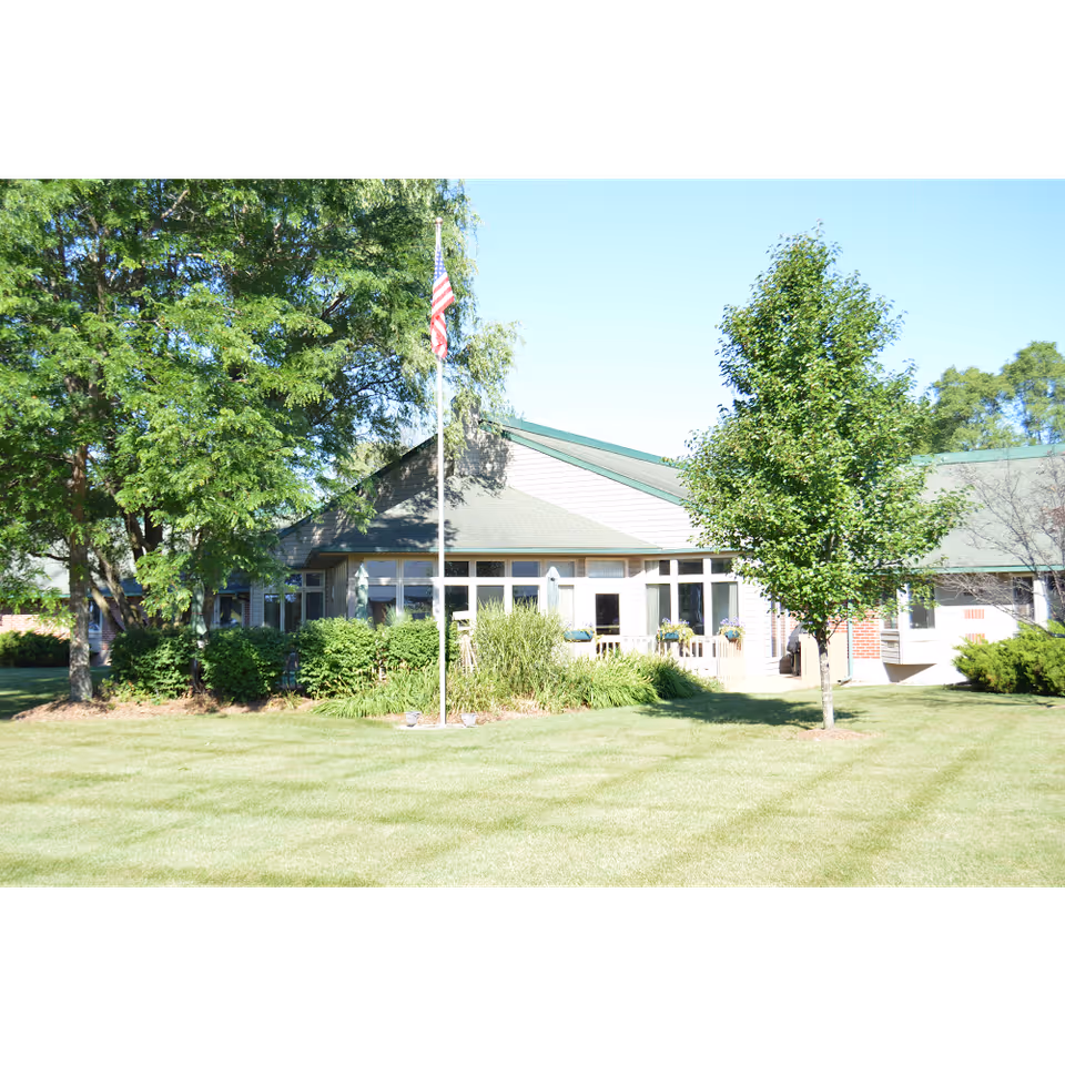 Exterior view of Bishop Hills Assisted Living facility showing a single-story building with large windows, surrounded by green trees, bushes, and a well-maintained lawn. An American flag is displayed on a flagpole in front of the building under a clear blue sky.