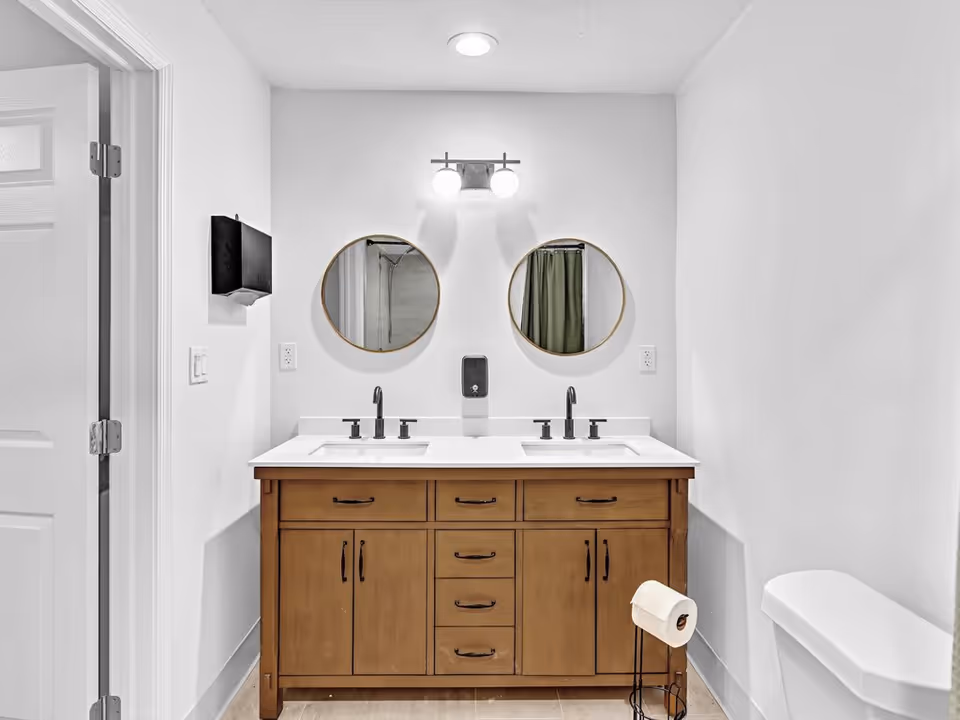 Bathroom with a wooden double-sink vanity, two round mirrors, overhead light, and a toilet.