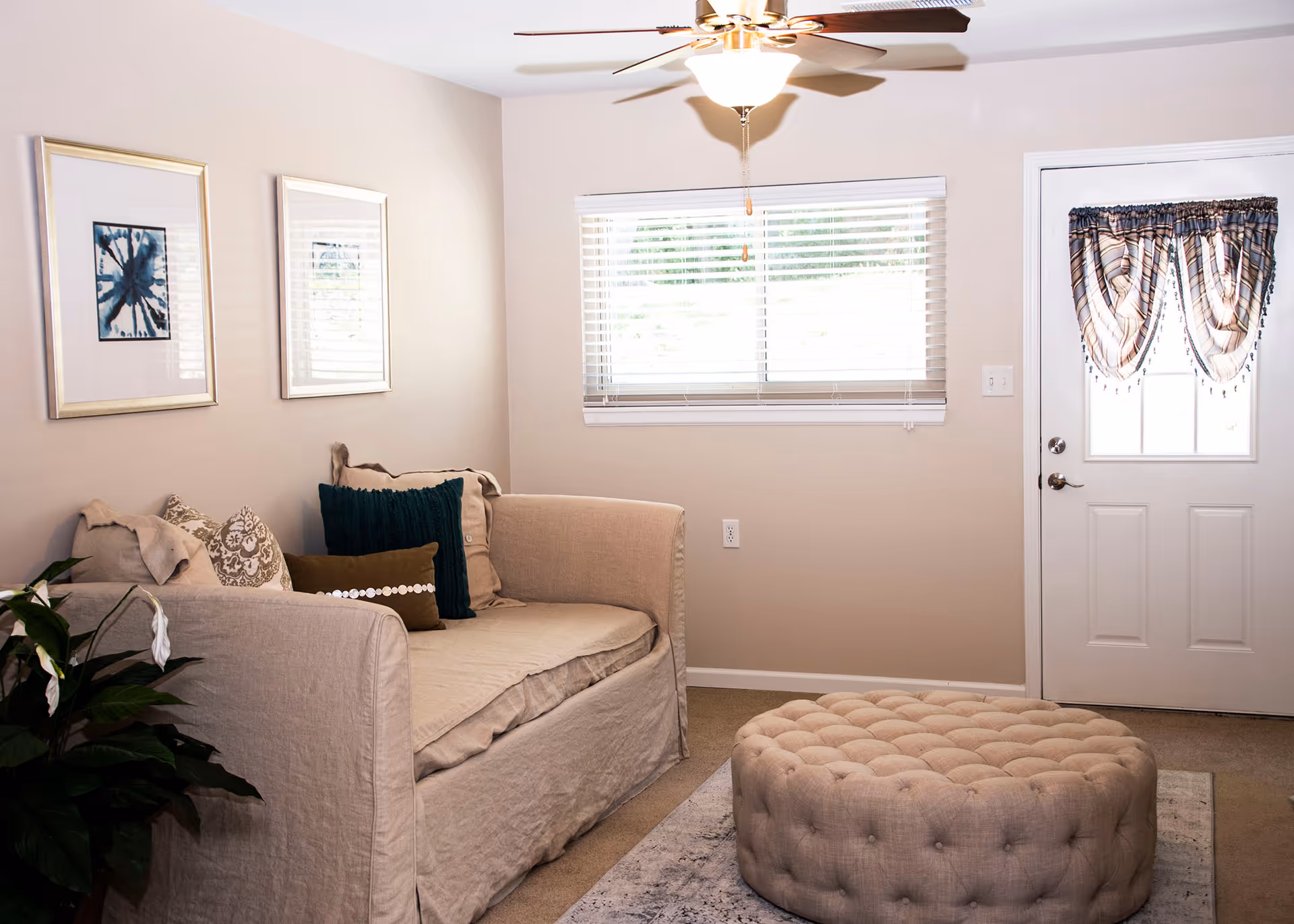 A cozy living room with a beige sofa adorned with multiple cushions, a round tufted ottoman in front, a ceiling fan with a light, two framed abstract artworks on the wall, a window with blinds, a door with a decorative curtain, and a green plant in the corner.