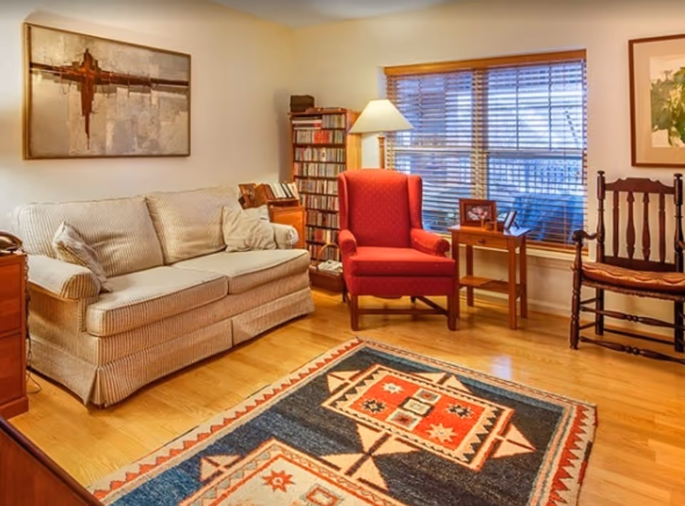 A cozy living room with a beige couch, a red armchair, a wooden chair with a cushion, a wooden side table with framed photos, a floor lamp, a bookshelf filled with CDs, and a large window with wooden blinds. The floor has a colorful patterned rug, and there are two framed artworks on the walls.