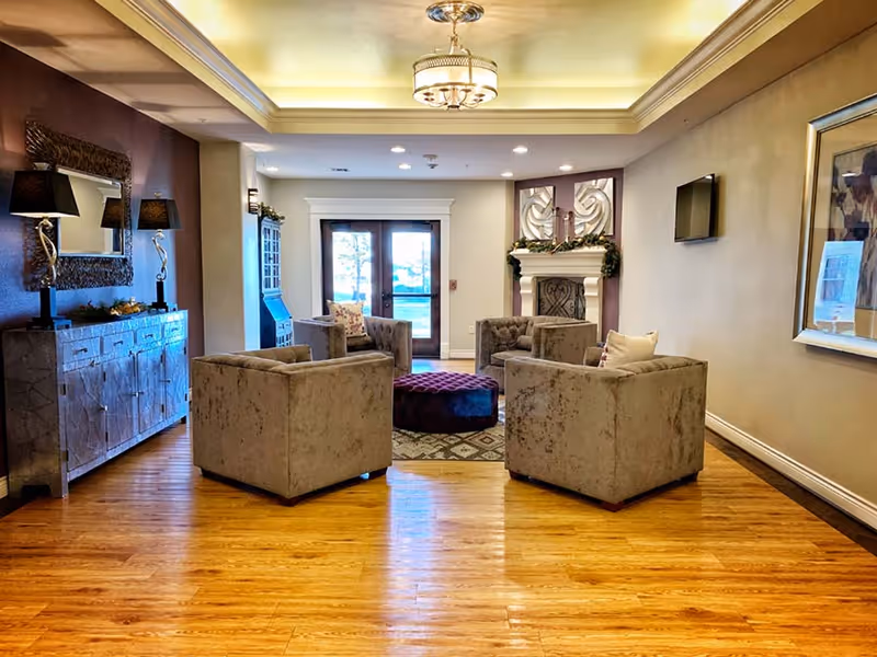 A cozy communal lounge with four tufted armchairs arranged around a round ottoman, a fireplace, hardwood floors and decorative ceiling lighting.