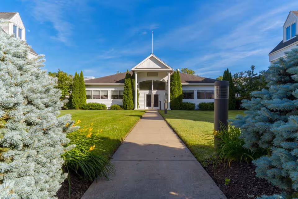 Front exterior view of a single-story building with a peaked roof and a central entrance, surrounded by green lawns, shrubs, and trees under a blue sky.