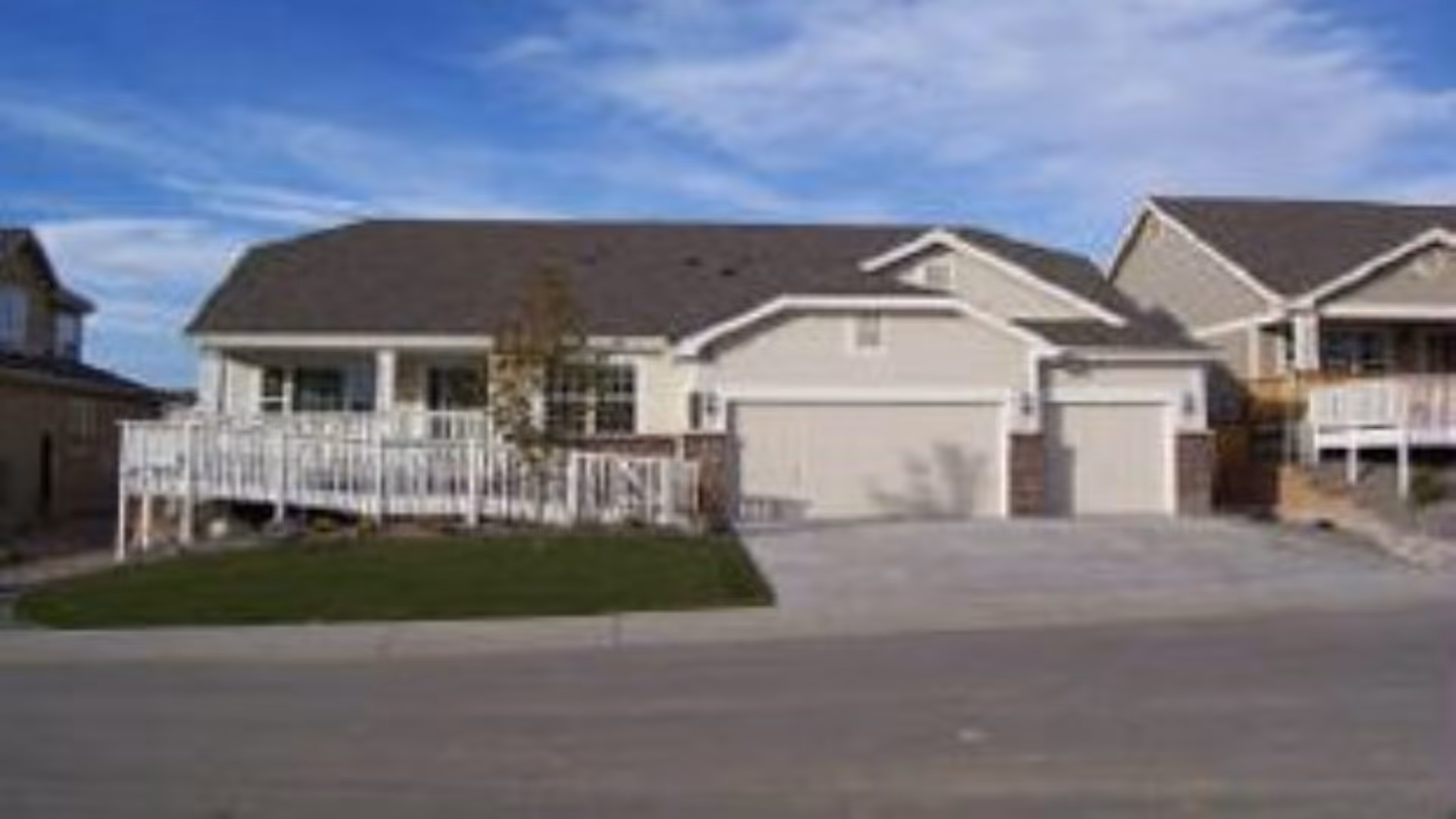 Front exterior view of a single-story residential building with a large driveway, a two-car garage, a small porch with white railings, and a well-maintained lawn under a partly cloudy blue sky.