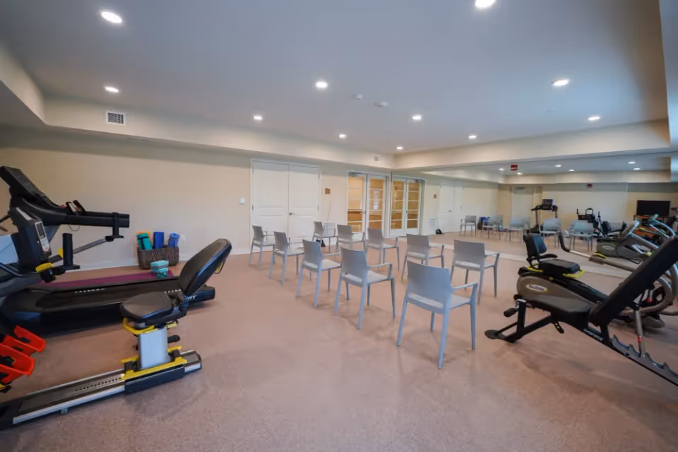 A fitness room with exercise equipment including treadmills, stationary bikes, and rowing machines. Several chairs are arranged in rows in the center of the room. The room has beige walls, a large mirror on one side, and recessed ceiling lights.