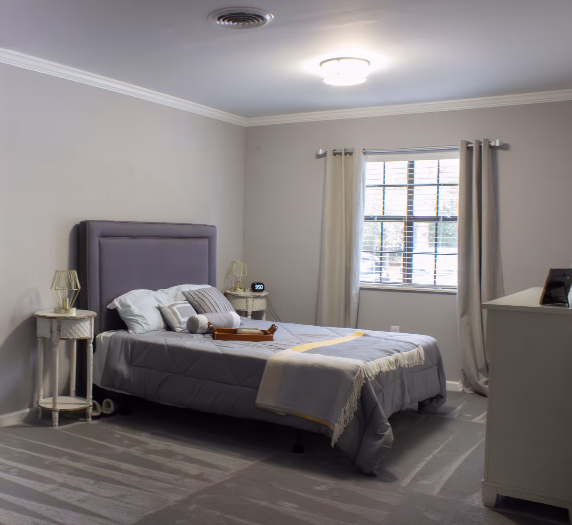 A neatly made bed with gray bedding and multiple pillows in a softly lit bedroom. The room features light gray walls, a window with beige curtains, two small round bedside tables with lamps, a digital alarm clock, and a white dresser with a framed photo on top.