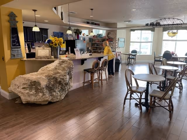 Interior view of a dining area in a senior living facility with wooden floors, round tables with chairs, a counter with bar stools, and a large decorative rock near the counter. A person in a yellow shirt is standing behind the counter. The room is well-lit with large windows and pendant lights.