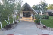 Front entrance of a senior living facility with a peaked canopy, a paved walkway, and landscaped beds with birch trees and lawn.