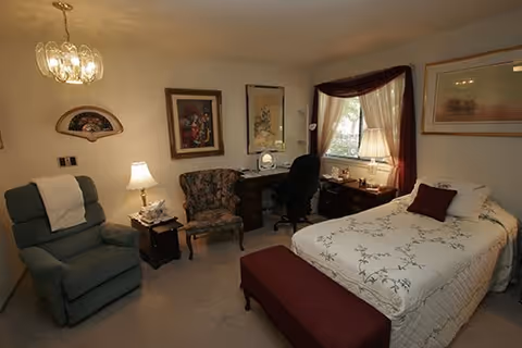 A cozy senior living bedroom featuring a bed with white floral bedding and a burgundy pillow, a matching burgundy bench at the foot of the bed, a green recliner chair with a white towel draped over it, a floral upholstered armchair, a wooden desk with a black office chair, a table lamp, framed artwork on the walls, and a window with burgundy curtains allowing natural light into the room.