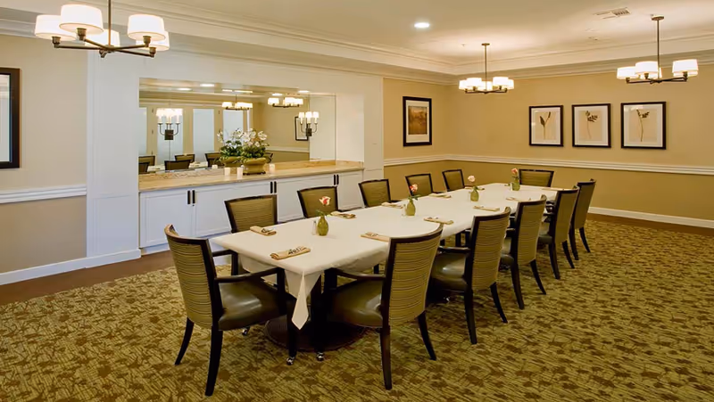 A well-lit dining room with a long rectangular table covered with a white tablecloth. The table is set with napkins and small vases holding flowers. There are twelve chairs around the table. The room has beige walls with white trim, framed artwork, and multiple ceiling light fixtures. A large mirror and a countertop with decorative plants are visible on one wall.