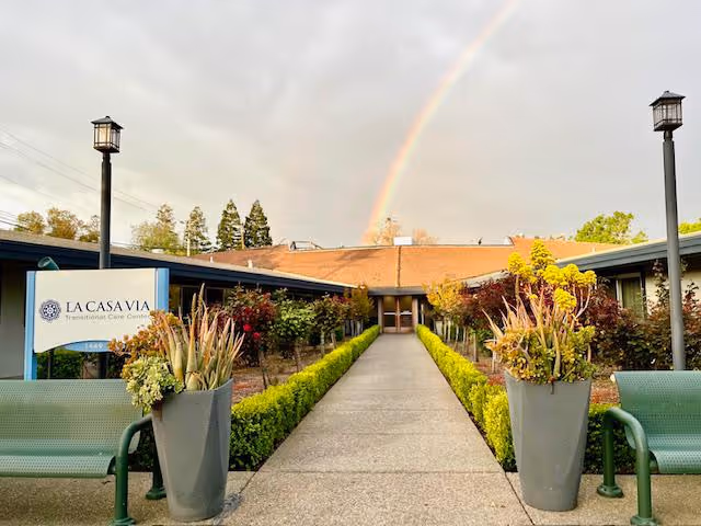 Front entrance courtyard of La Casa Via Transitional Care Center with a walkway flanked by planters and benches and a rainbow overhead.
