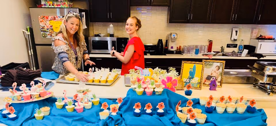 Two women standing behind a kitchen counter preparing and arranging decorated cupcakes with tropical-themed toppers. The kitchen has dark cabinets, a microwave, and a refrigerator with colorful magnets and notes. The counter is covered with a blue cloth displaying multiple cupcakes with various decorations.
