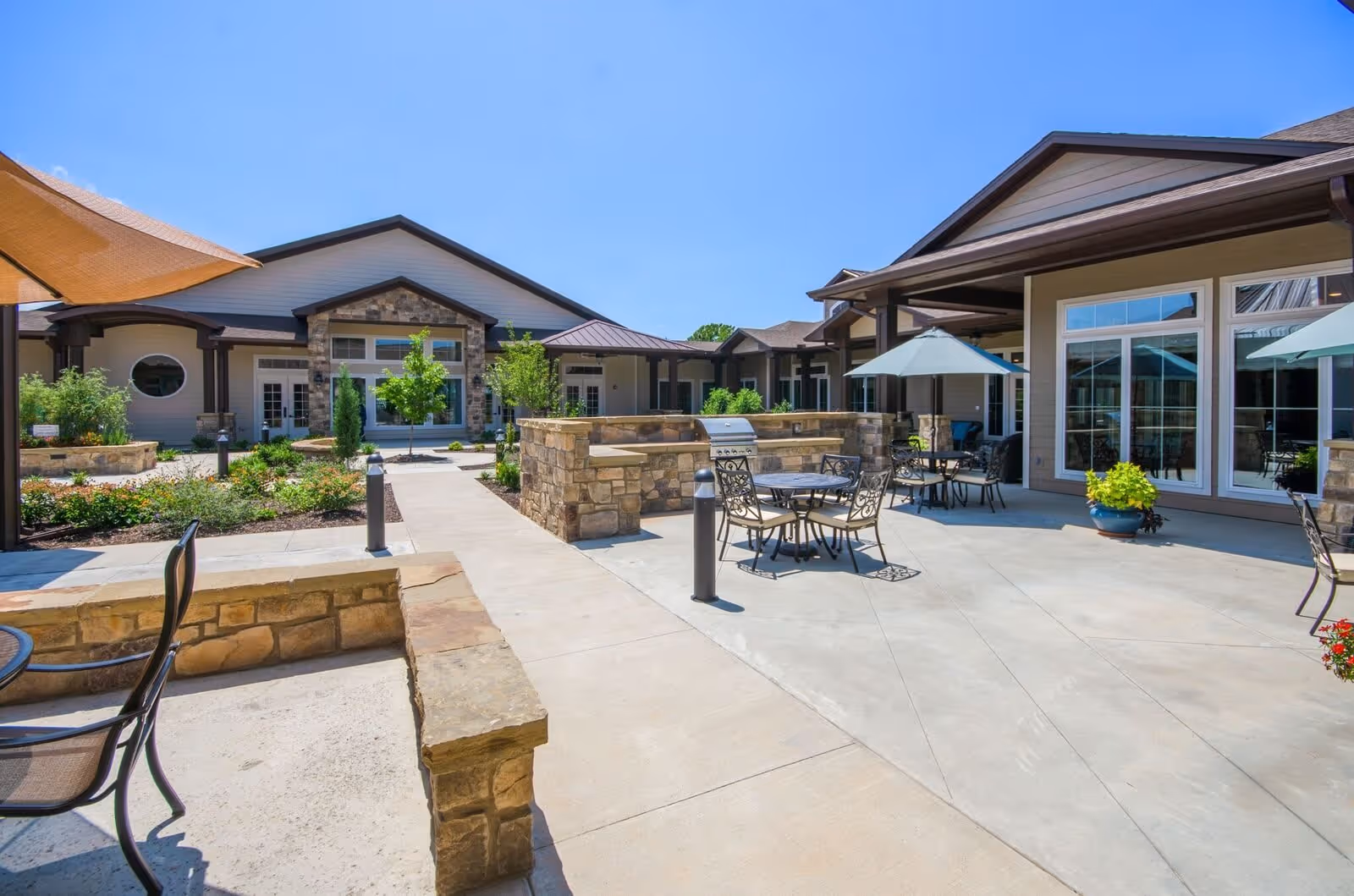 Outdoor patio area at Grand Brook Memory Care of Zionsville featuring stone walls, metal tables and chairs with umbrellas, a built-in grill, and landscaped garden beds under a clear blue sky.