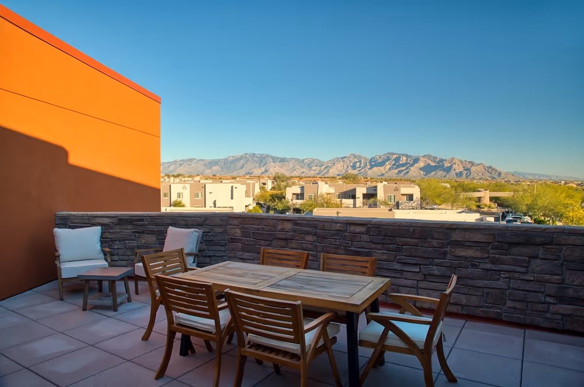 Outdoor patio area with a wooden table and six chairs, two additional cushioned chairs with a small side table, stone wall railing, and a view of residential buildings and mountains under a clear blue sky.