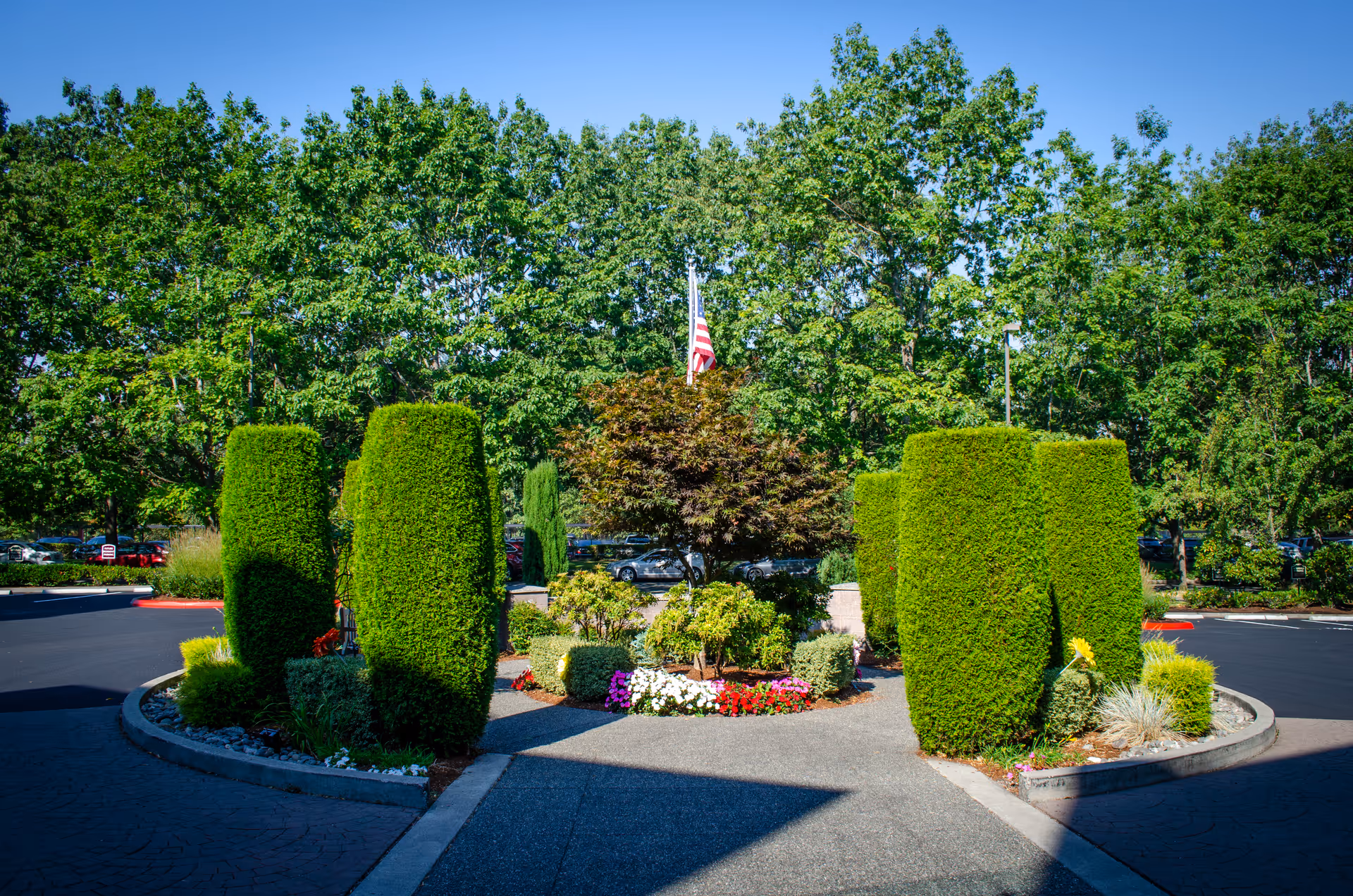 A landscaped outdoor area with neatly trimmed tall green bushes and various colorful flowers arranged in a circular garden bed. Behind the garden, there are large green trees and an American flag on a flagpole. The area is surrounded by a paved driveway and parking lot with some parked cars visible in the background under a clear blue sky.