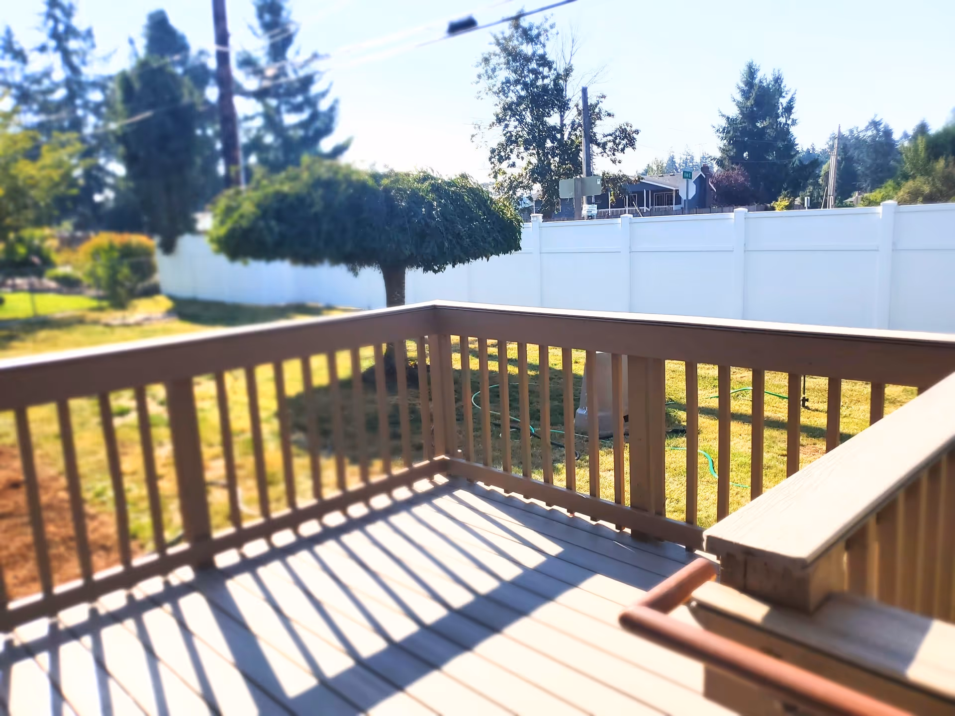 View from a wooden deck overlooking a fenced backyard with green grass, a small tree, and other trees in the distance under a clear sky.