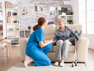 A healthcare worker in blue scrubs kneels beside an elderly woman sitting on a beige couch in a bright living room. The elderly woman has crutches resting against the couch and they are holding hands, smiling at each other. The room has wooden flooring, a white bookshelf with decorations, and large windows letting in natural light.