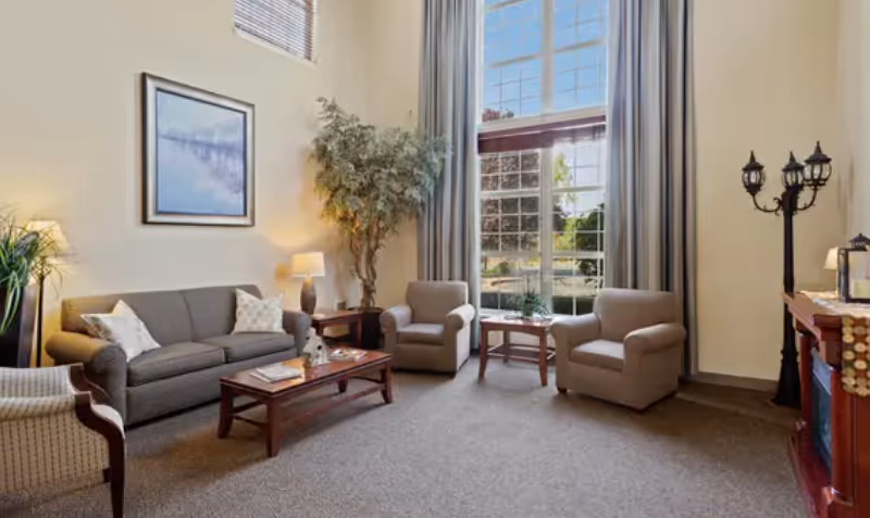 A cozy senior living facility common area with a gray sofa, two beige armchairs, a wooden coffee table, a side table with a lamp, a large indoor plant, and a tall window with curtains letting in natural light.