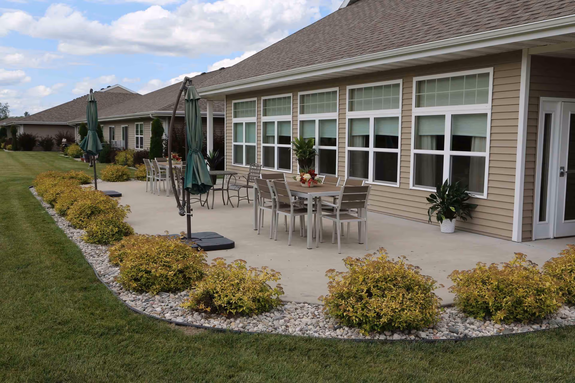 Outdoor patio area of a senior living facility with multiple tables and chairs, closed green umbrellas, potted plants, and a well-maintained lawn with bushes and decorative rocks along the edge of the concrete patio.