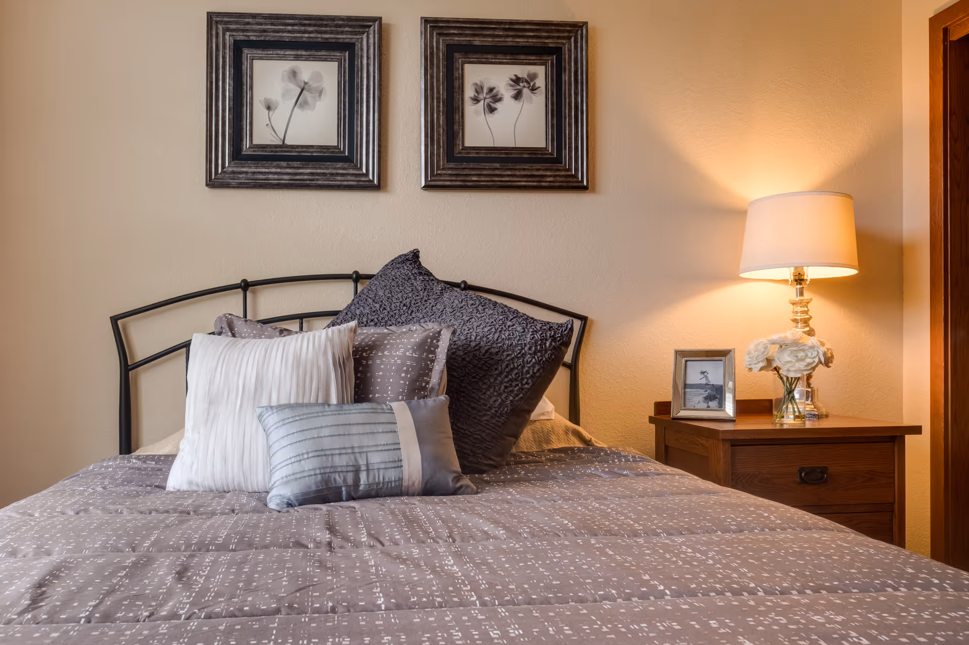 Neatly made bed with decorative pillows, framed artwork above, and a bedside table with a lamp and vase of flowers.