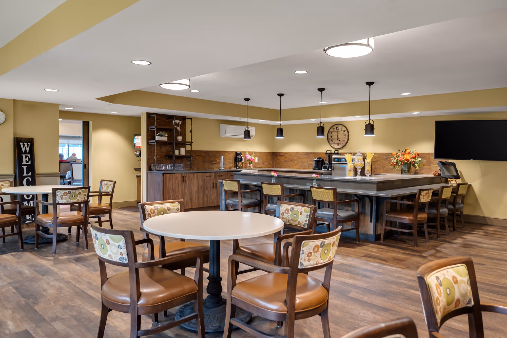 A spacious dining area with round tables and cushioned chairs featuring patterned backs. The room has wooden flooring and warm yellow walls. There is a large counter with bar stools, pendant lights hanging above, and a wall clock. A flat-screen TV is mounted on the wall, and there are decorative flowers on the counter. A welcome sign is visible near the entrance.