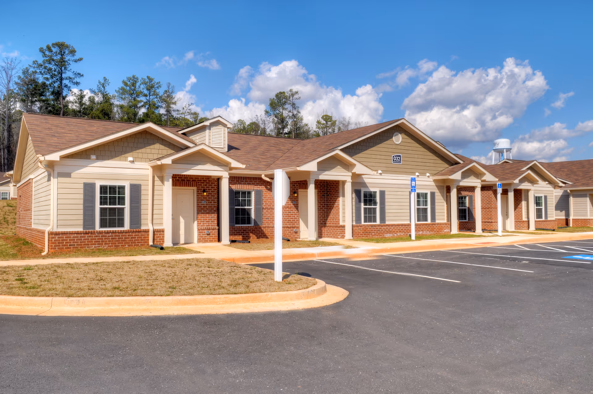 Single-story brick-and-siding senior living building with covered entrances, a nearby water tower, and an empty parking lot under a blue sky.