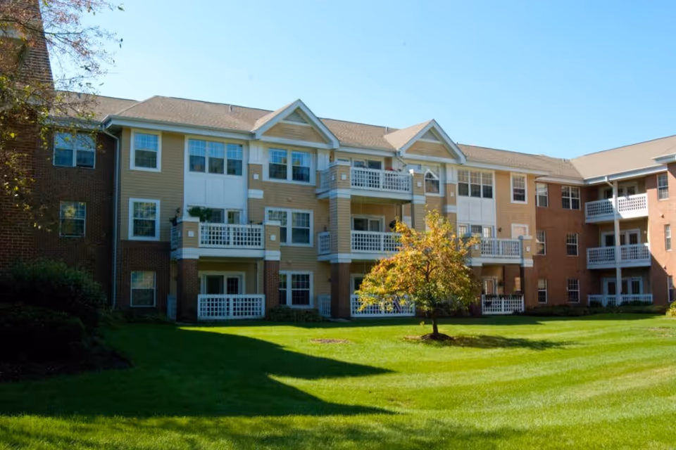 Exterior view of a multi-story residential building with balconies, surrounded by a well-maintained green lawn and a small tree in the center under a clear blue sky.