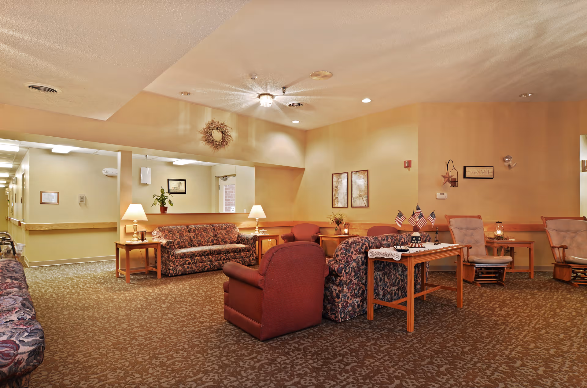 A cozy senior living facility common area with patterned sofas and armchairs arranged around a small wooden table decorated with miniature American flags. The room has warm beige walls, carpeted flooring, framed artwork, and soft lighting from table lamps and ceiling fixtures. A hallway is visible to the left.