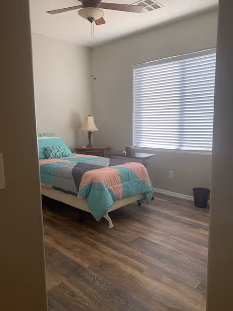A small bedroom with a single bed covered in a colorful quilt with shades of teal, pink, and gray. There is a wooden nightstand next to the bed with a lamp on it. A window with closed blinds lets in natural light. The floor is wood with a medium brown finish, and a ceiling fan is visible above.