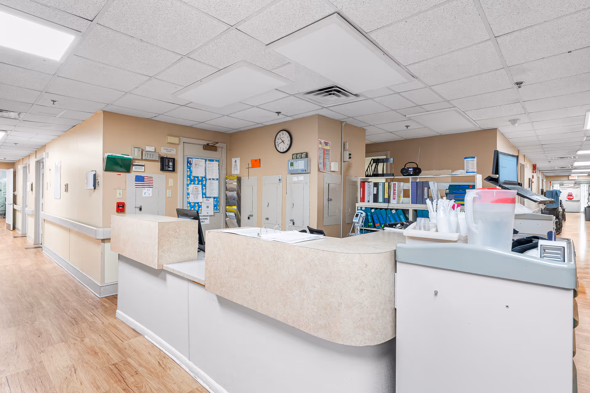 Interior view of a nursing station in a senior living facility with a curved counter, office supplies, computer monitors, shelves with binders, and a hallway extending to the left and right.