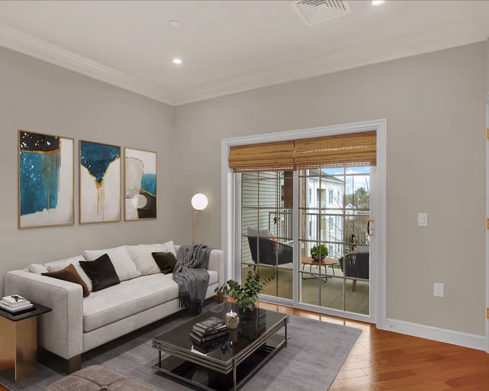 A modern living room with a light gray sofa adorned with multiple cushions and a gray throw blanket. Above the sofa are three abstract art pieces with blue, gold, and white tones. A black glass coffee table with books, a candle, and a small plant sits on a gray rug. To the right, there is a sliding glass door with bamboo blinds, leading to a balcony with two black chairs and a small round table with a plant. The room has light gray walls and wooden flooring.