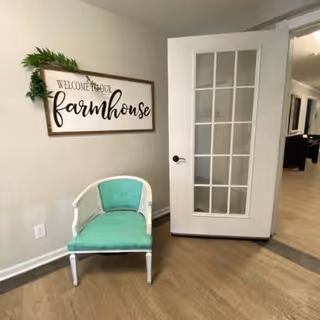 A small corner of an interior room featuring a white chair with a green cushion placed against a beige wall. Above the chair is a decorative sign that reads 'Welcome to our farmhouse' with some green foliage attached to the top left corner of the sign. To the right, there is a white door with glass panes that is partially open, revealing a hallway with light wood flooring and additional rooms in the background.