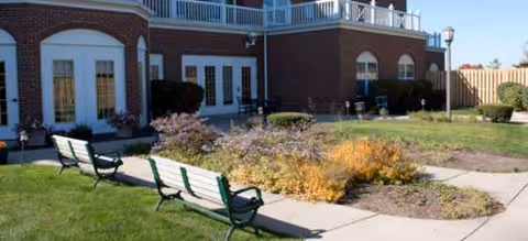 Brick senior living facility exterior with arched windows, benches, and manicured landscaping.