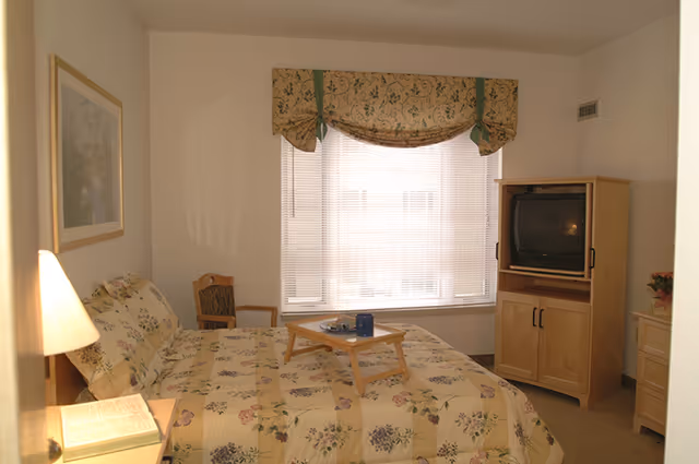 Sunlit bedroom with a floral-patterned bed and breakfast tray, window with blinds and valance, wooden TV cabinet and nightstand with a lamp.