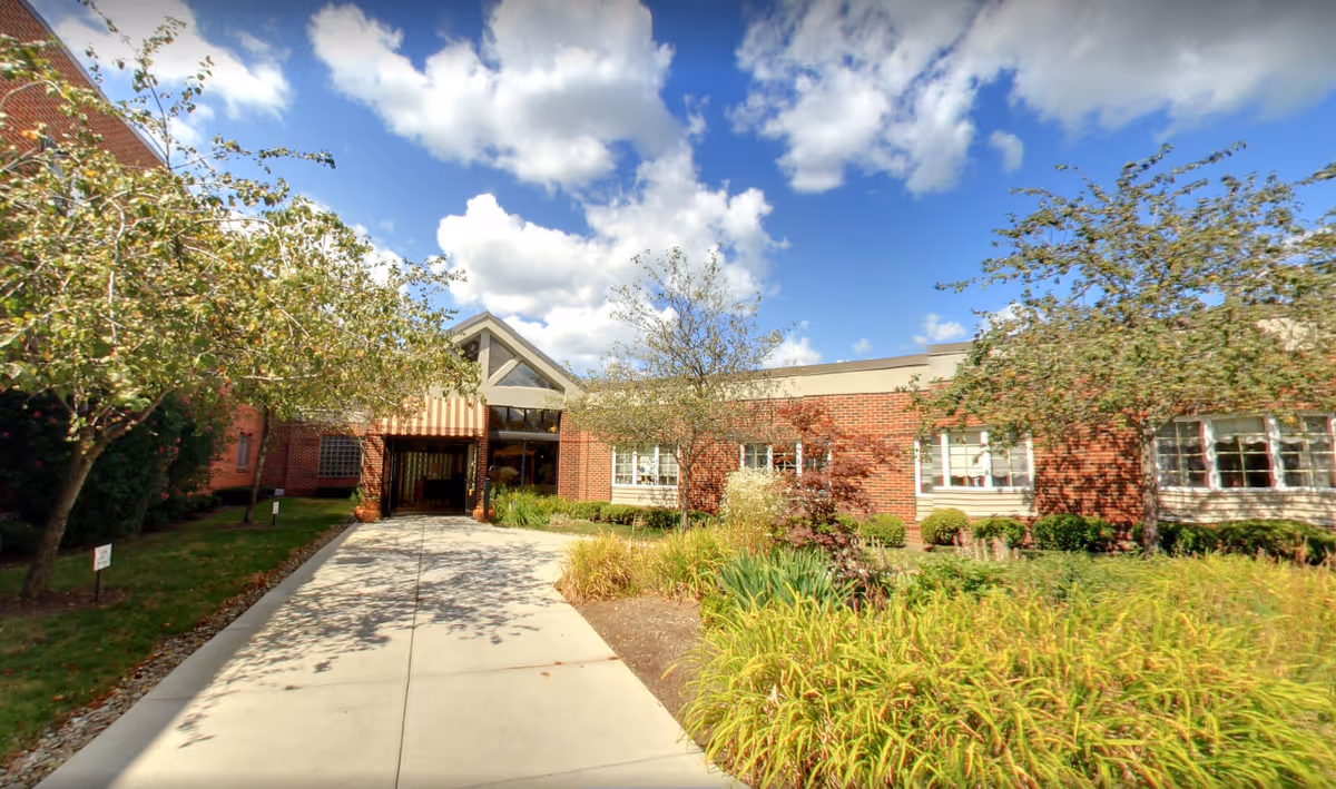 Entrance to a brick building with a concrete walkway leading up to a covered entryway. The building is surrounded by green shrubs, trees, and landscaped plants under a partly cloudy blue sky.