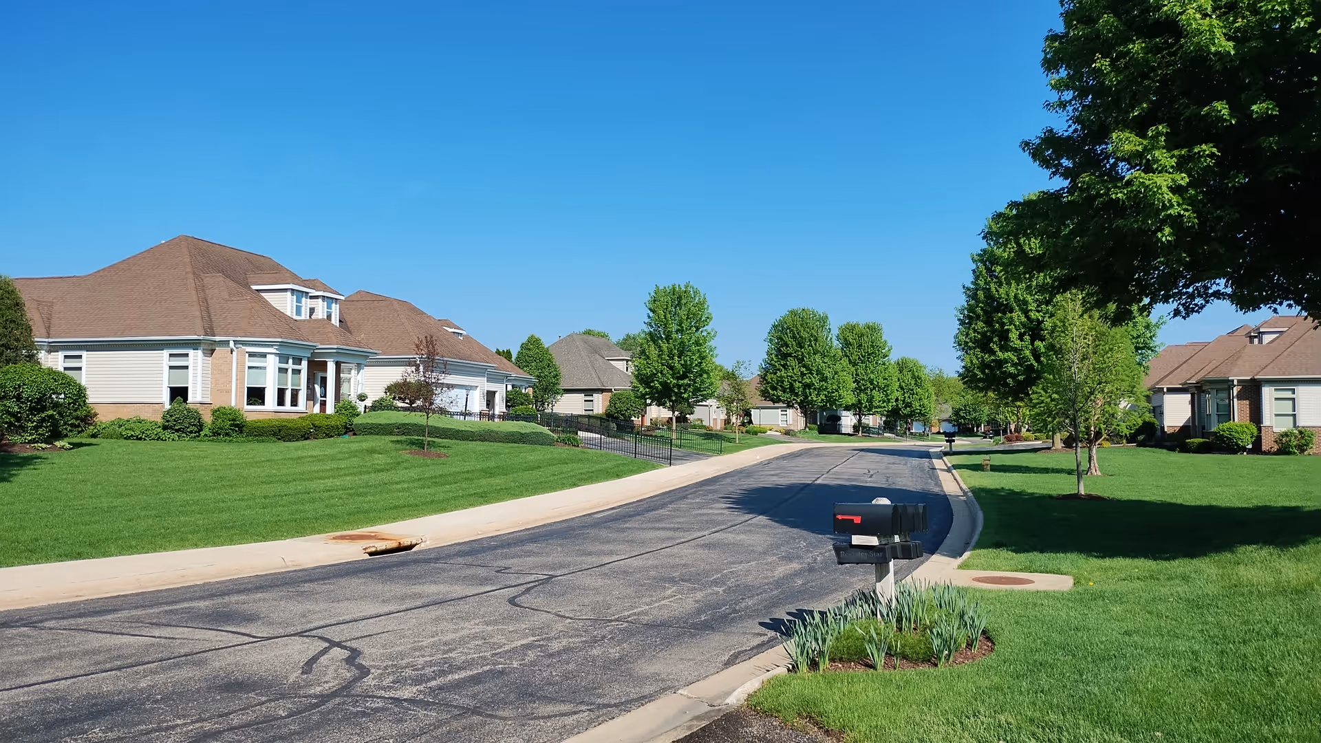A quiet residential street in a senior living community with well-maintained green lawns, trees, and single-story houses with brown roofs under a clear blue sky.