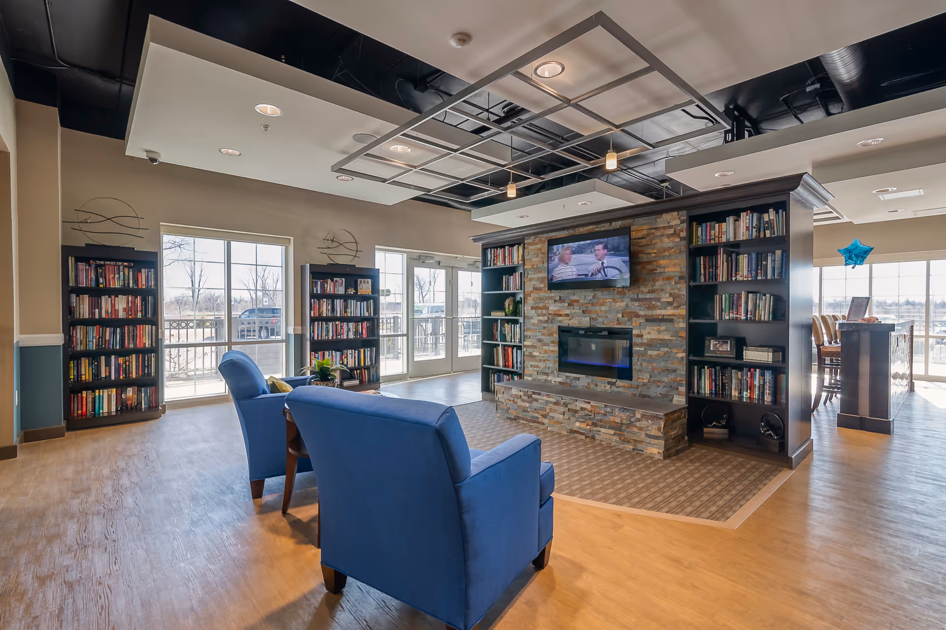 A cozy living room area with two blue armchairs facing a stone fireplace with a mounted flat-screen TV above it. The fireplace is flanked by built-in bookshelves filled with books. Large windows and glass doors let in natural light and provide a view of the outdoors. The ceiling has modern light fixtures and an exposed black ceiling design.