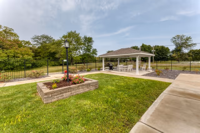 Outdoor area at High Point Residence Centralia featuring a green lawn with a raised flower bed containing blooming flowers and a black lamp post. In the background, there is a white gazebo with a seating area including white chairs and a table, surrounded by a black metal fence and trees under a partly cloudy sky.