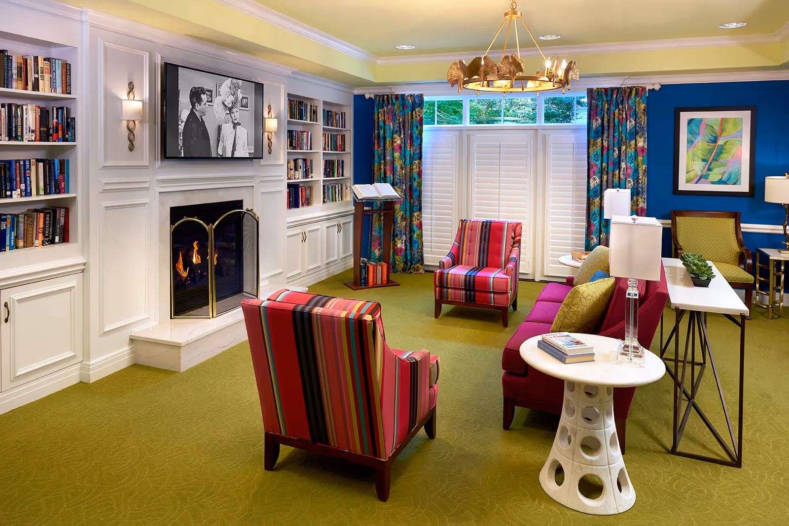 A colorful assisted-living common room with a fireplace and built-in bookshelves, striped armchairs, a sofa, and a large window with shutters.
