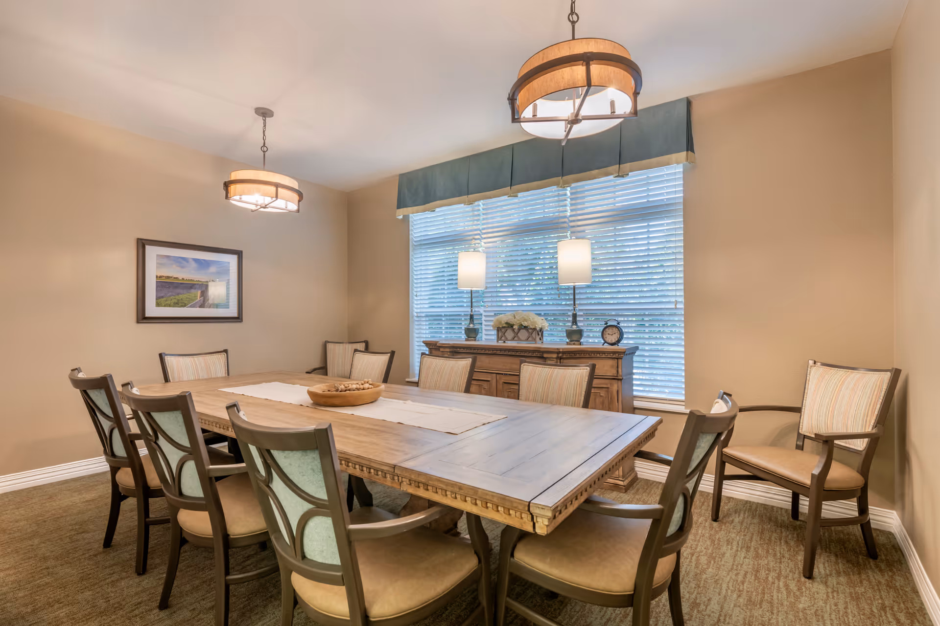A dining room with a large wooden table surrounded by eight cushioned chairs. Two pendant lights hang from the ceiling above the table. A sideboard with two table lamps, a flower arrangement, and a clock is positioned against a window with blinds and a blue valance. The walls are painted beige, and there is a framed landscape picture on one wall.