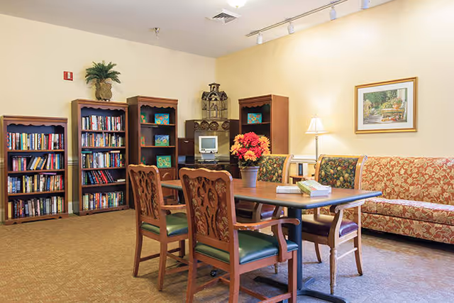 A cozy common area with a wooden table surrounded by four chairs with green cushions. Behind the table, there is a patterned sofa and a side table with a lamp and a vase of red and orange flowers. On the left side, there are three bookshelves filled with books and decorative items. A computer is placed on a small desk between the bookshelves. The walls are painted light yellow, and a framed picture hangs above the sofa.