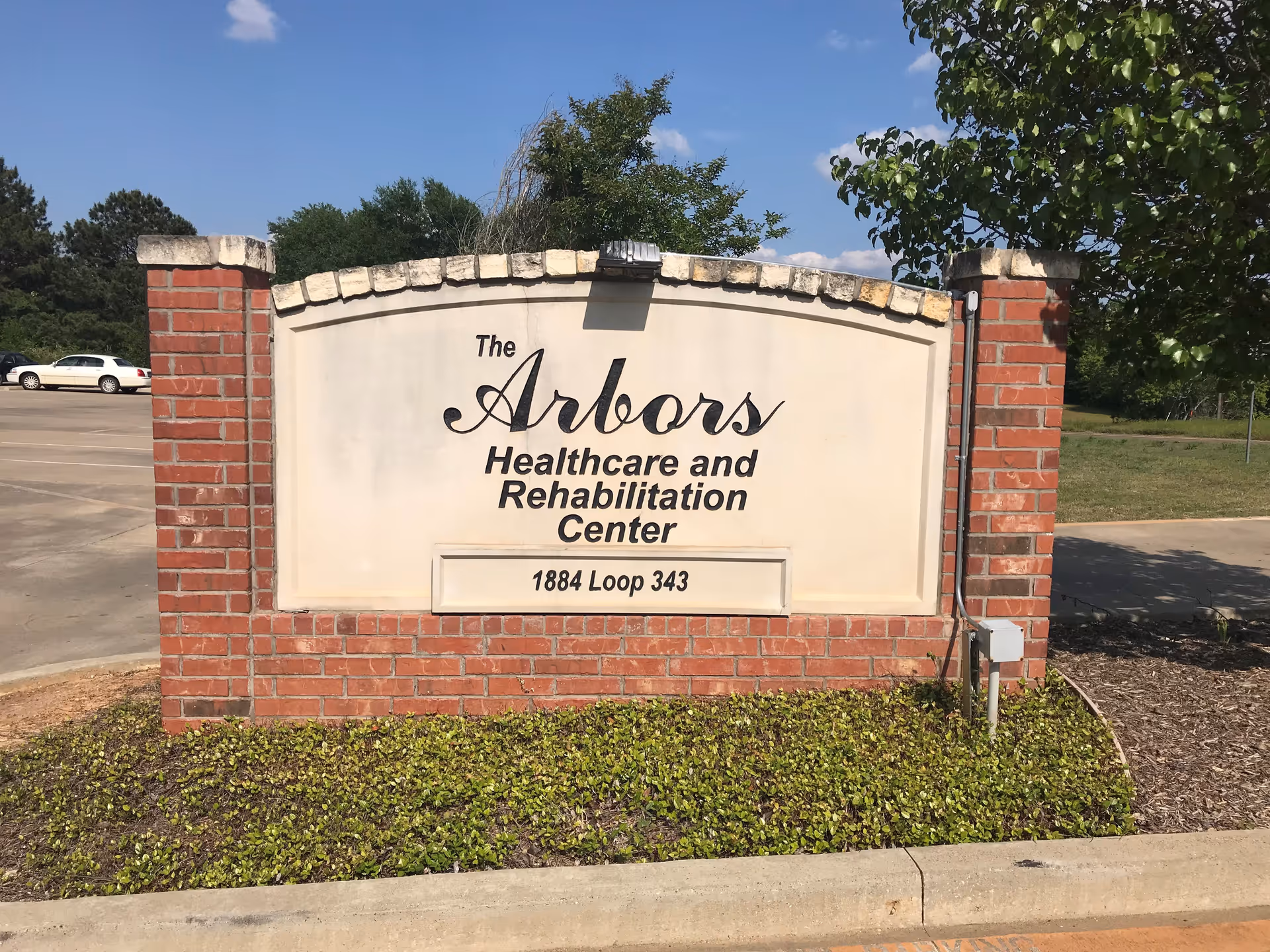 A brick and stone sign for The Arbors Healthcare and Rehabilitation Center located at 1884 Loop 343, with a parking lot and trees in the background under a clear blue sky.
