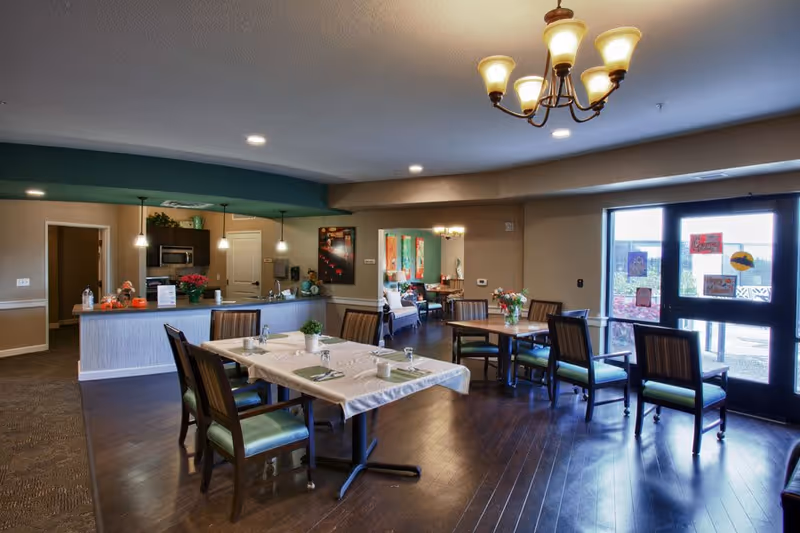 A dining area in a senior living facility with several tables and chairs arranged neatly. The tables are set with placemats, napkins, and glassware. In the background, there is a kitchen area with a counter, pendant lights, and a microwave. The room has wooden flooring, warm lighting from ceiling fixtures, and a glass door leading outside with decorations on it.