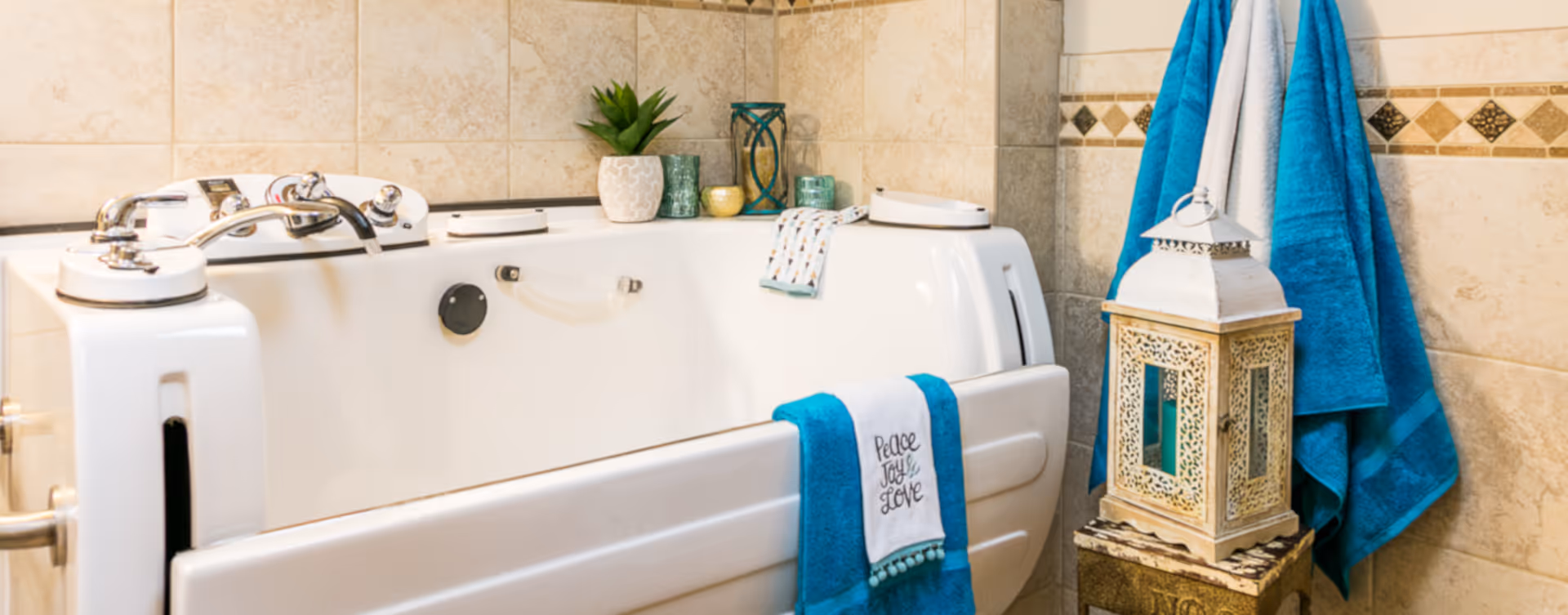 A white walk-in bathtub in a tiled bathroom with silver faucets. A blue towel with the words 'Peace Joy Love' hangs over the tub. There are decorative items including a small plant, candles, and a lantern on a stand next to the tub. Blue towels hang on hooks on the wall.