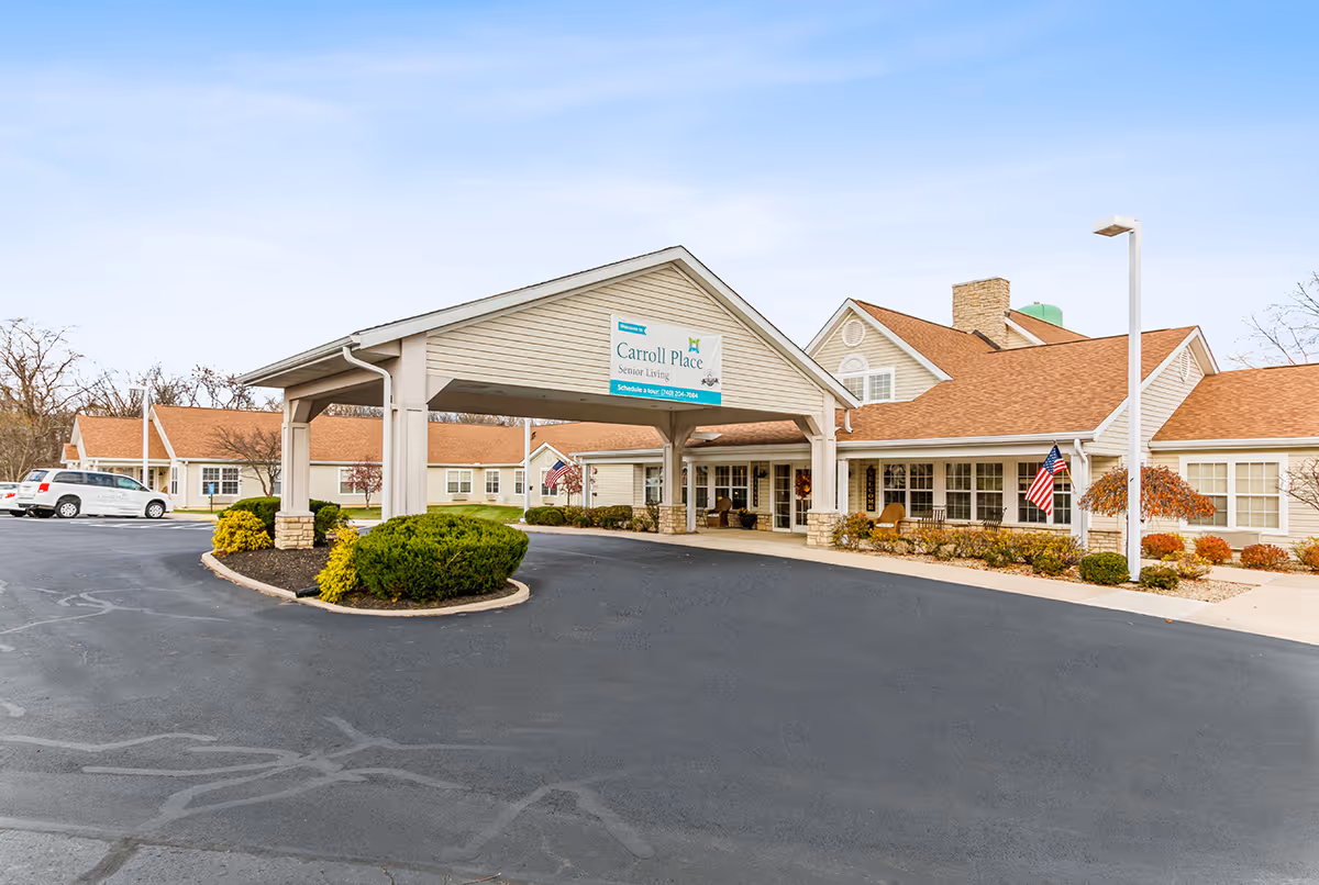 Front entrance of Carroll Place senior living facility with a covered drive-through portico, landscaped beds, and American flags.