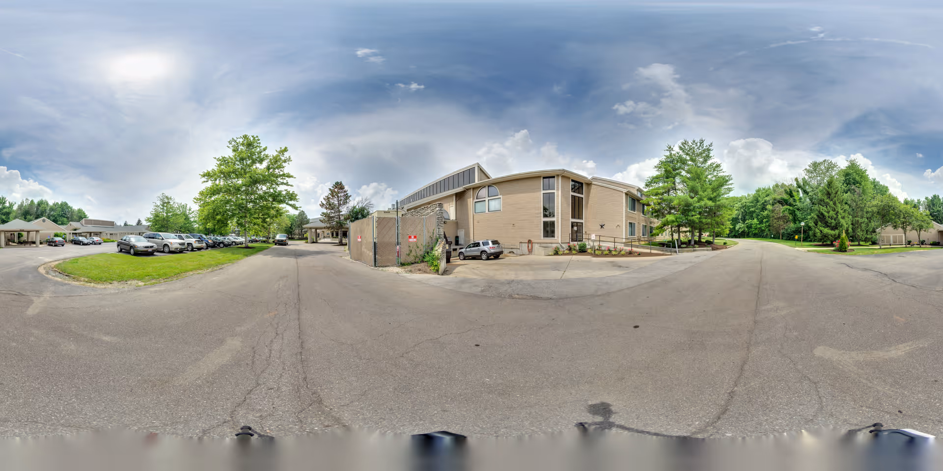 Exterior panoramic view of a retirement community building with a parking lot, parked cars, driveways, and surrounding trees under a cloudy sky.
