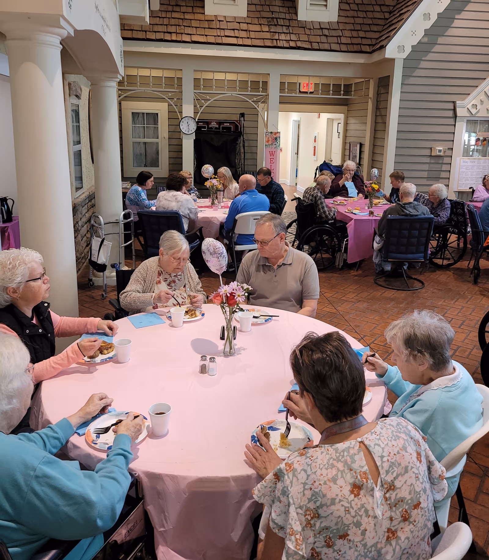 A group of elderly people sitting around tables covered with pink tablecloths, eating and socializing in a spacious indoor common area with high ceilings and decorative columns. The room has a warm, inviting atmosphere with flowers on the tables and a clock on the wall.