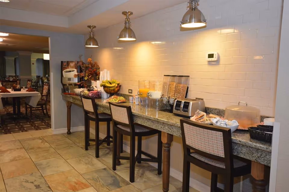 A breakfast buffet area with a granite countertop featuring cereal dispensers, a toaster, juice containers, fruit bowl, and covered food items. Three chairs are placed along the counter, and the area is lit by three hanging pendant lights. In the background, a dining room with tables and chairs is visible.
