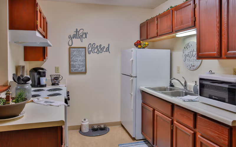 A small kitchen with wooden cabinets, a white refrigerator, a white stove with a coffee maker on top, a double sink, and a microwave. Wall decorations include the words 'gather' and 'blessed' and a framed sign with an inspirational message. There is a bowl with utensils and a plant on the counter.