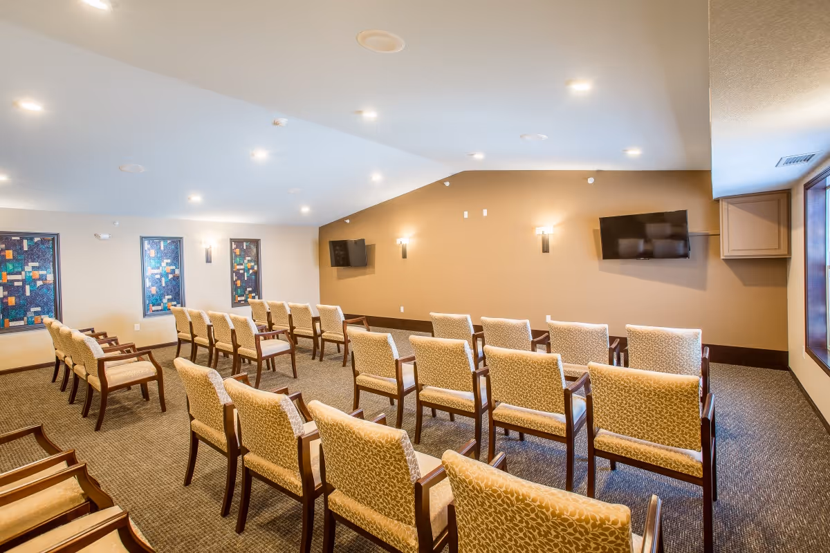 A well-lit room with rows of beige patterned chairs arranged facing two wall-mounted flat-screen TVs on a tan accent wall. The room has carpeted flooring, three decorative stained glass windows on one side, and recessed ceiling lights.