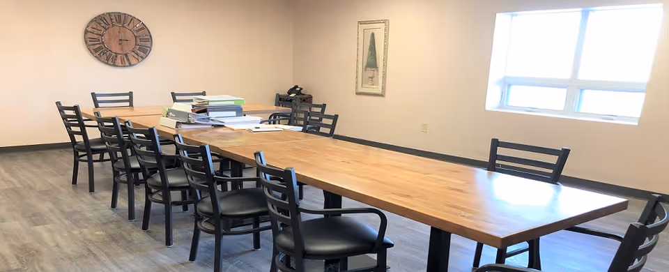 A long wooden conference table with black chairs arranged around it in a well-lit room. On the table, there are stacks of binders and papers. The room has light-colored walls, a large clock on one wall, a framed picture, and a window letting in natural light.